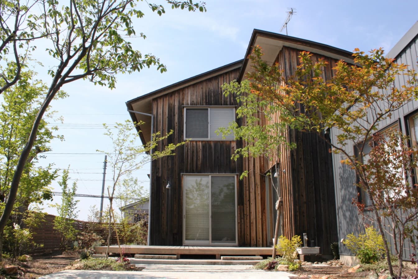 木の質感を活かした壁が印象的なダークブラウンのお家と、緑の植物が生える庭