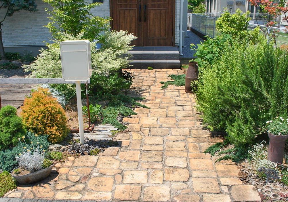 ベージュ系のレンガでできたアプローチと緑の植物が生えている、白いポストが印象的な庭