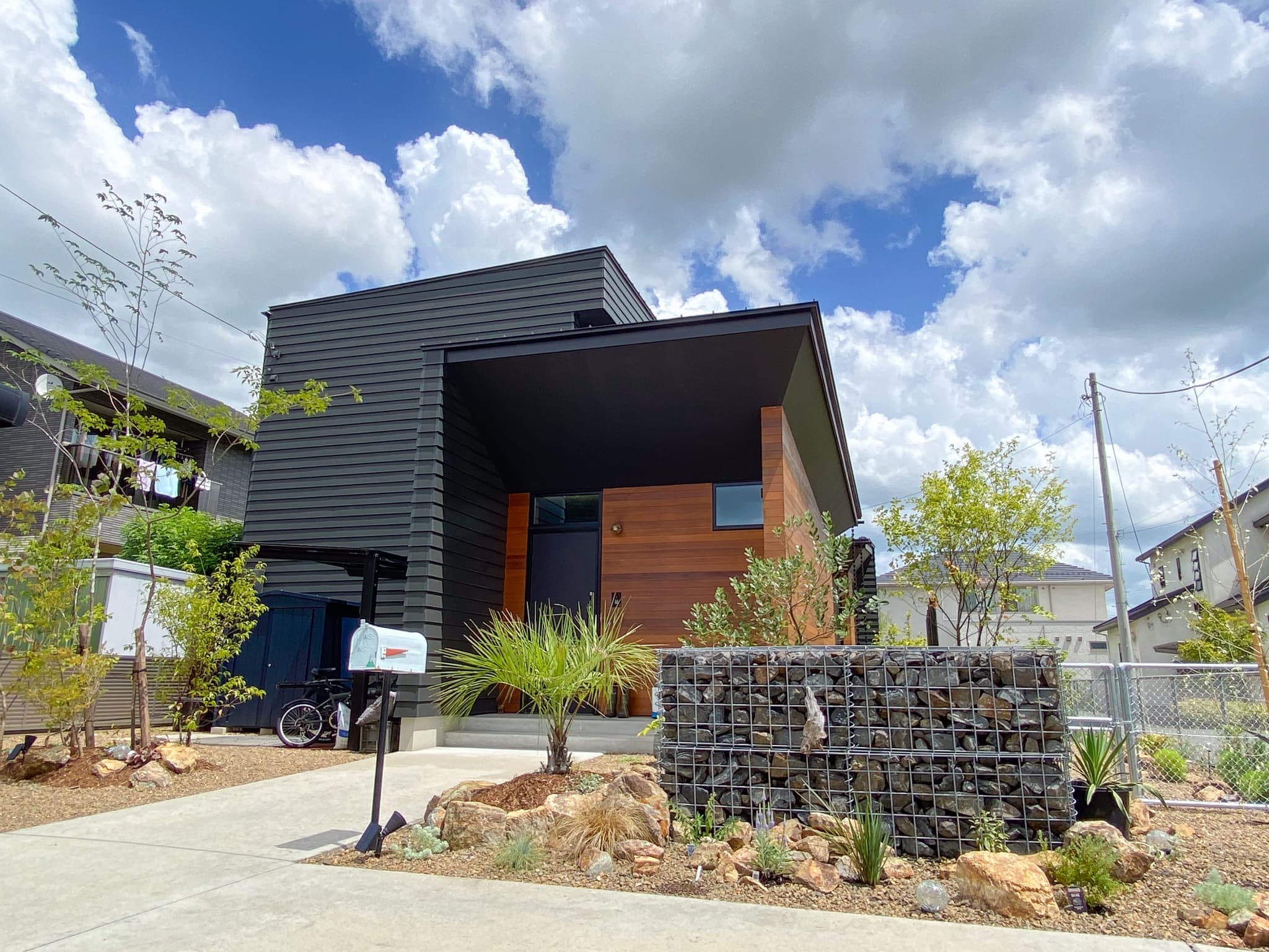 黒いモダンな建物。庭にはガビオンや植物、白いポストが設置されています