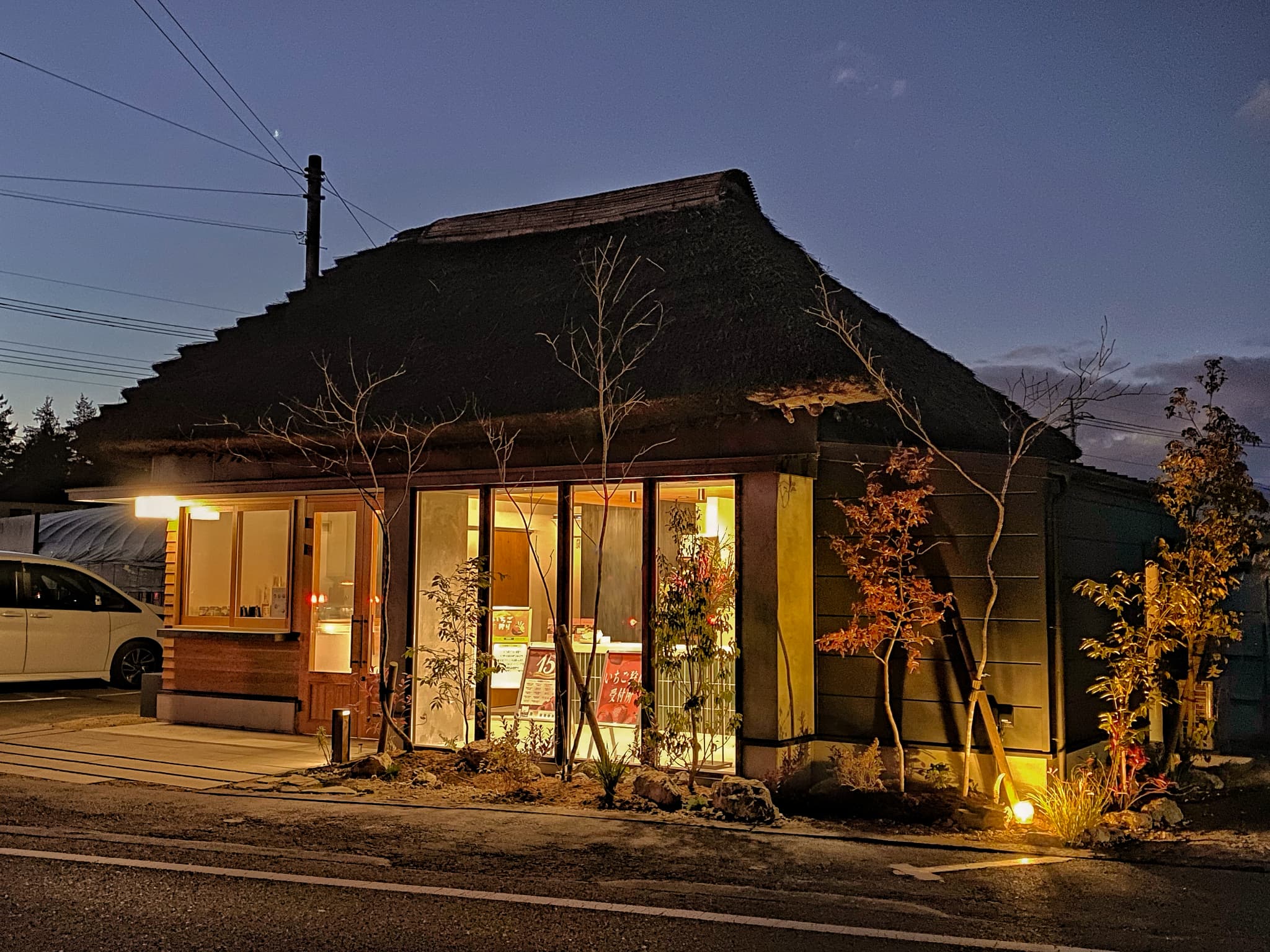 夜に黄色い温かみのある電気がついた茅葺き屋根が印象的な黒い建物の周辺に植物が生えている