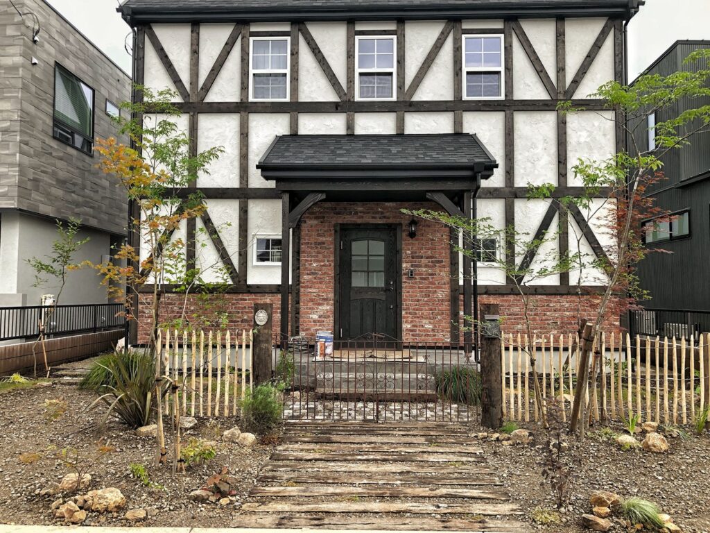 木枠と白壁が特徴の洋風住宅の外観。レンガづくりの玄関周りと小さな木製フェンス、砂利と若木が植えられたナチュラルな前庭が見える。