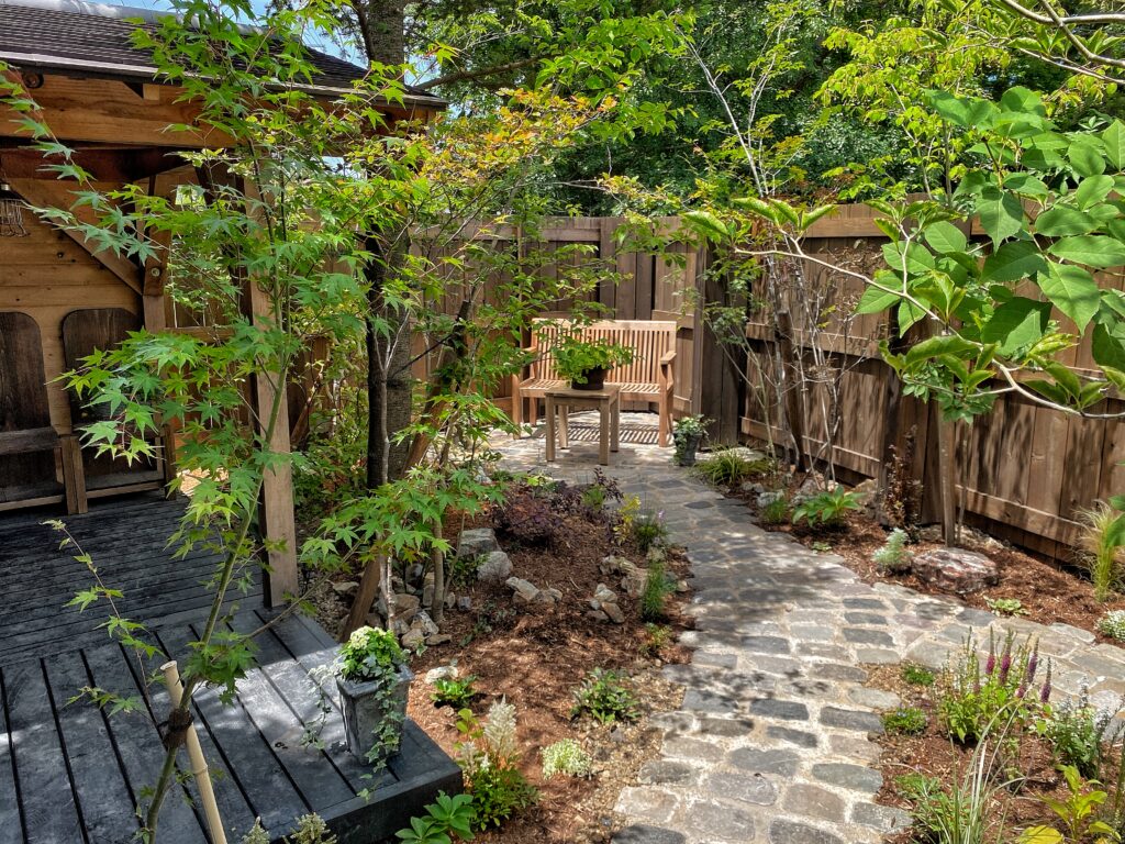 木製デッキと緑豊かな庭。石畳の小径が木々と草花に囲まれ、落ち着いた自然風のガーデンスペースが広がる。
