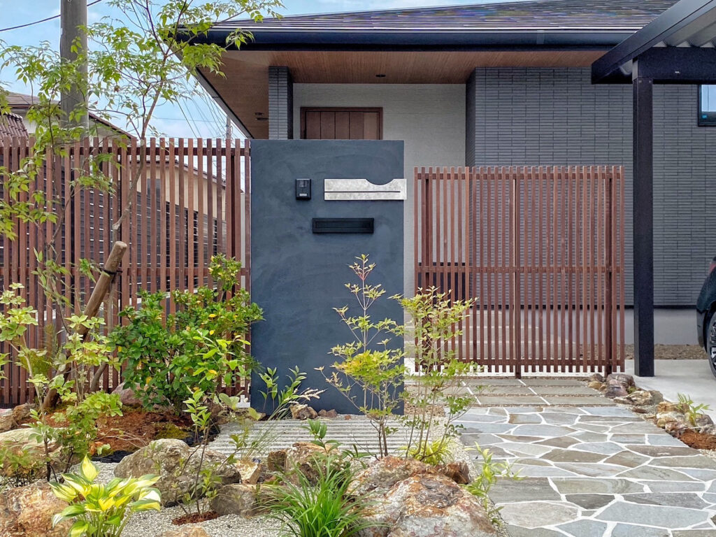 石張りのアプローチと黒い門柱、木製格子フェンスに囲まれたモダンな住宅の玄関周り。植栽が彩りを添える落ち着いた外構。